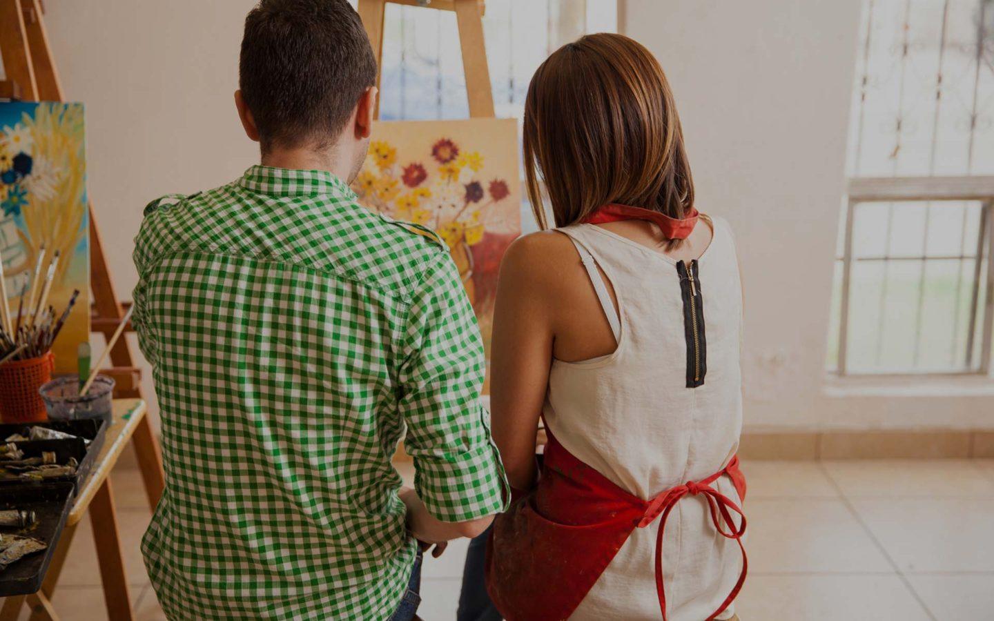 a man wearing a green shirt sits next to.a woman wearing a red apron, both are painting on an easel